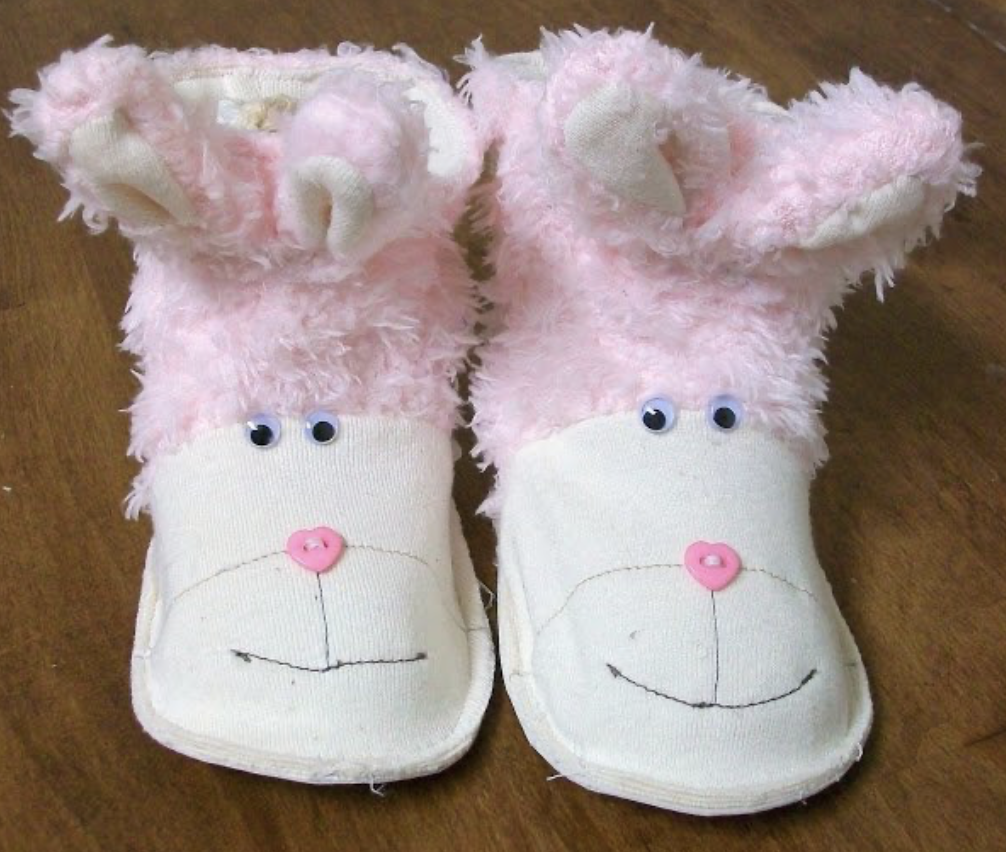 Pink and white children's slippers with fluffy material showcase a cute bunny design, complete with ears, eyes, and a button nose. They rest charmingly on a wooden surface, reminiscent of adorable sewing patterns brought to life.