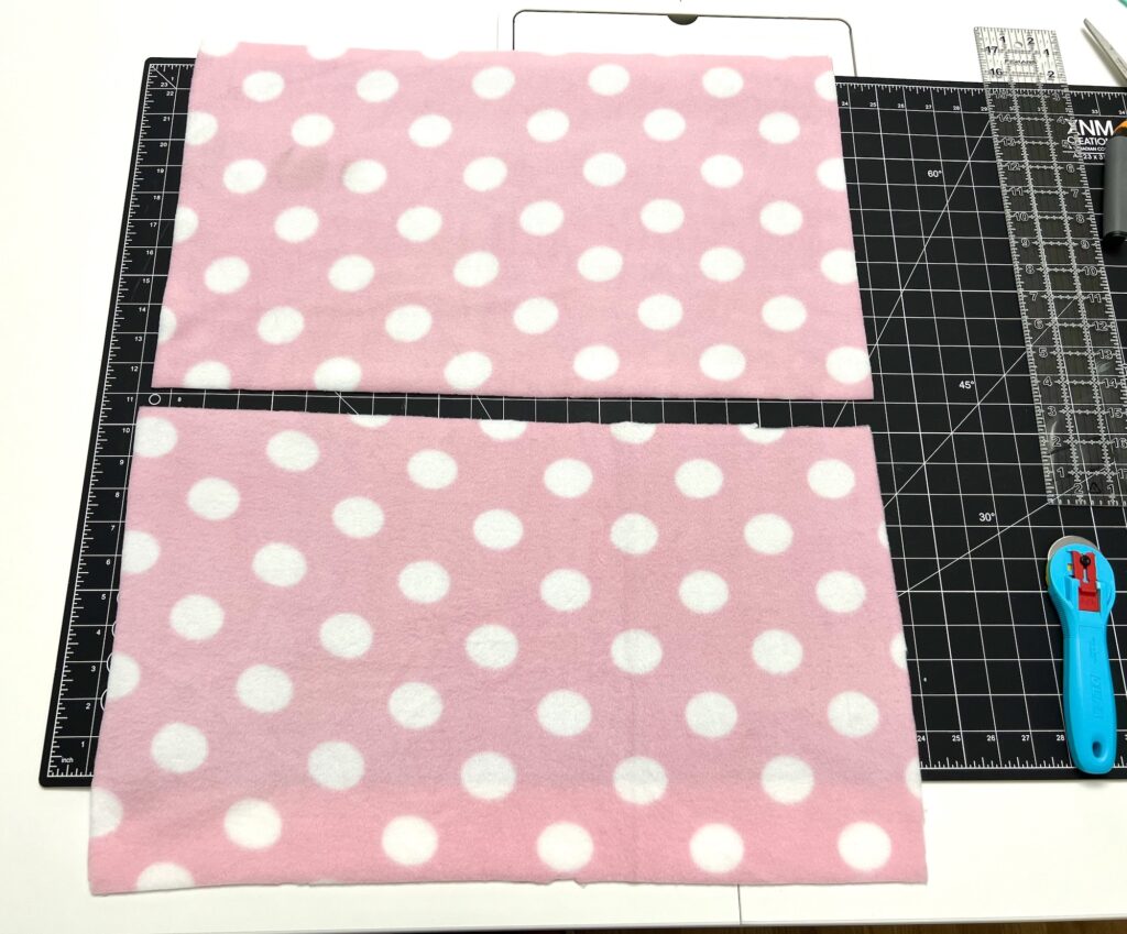 Two pieces of pink fabric with white polka dots rest on a black cutting mat, perfect for crafting a cozy fleece hat. A rotary cutter and clear measuring ruler accompany the setup, making the creative process seamless and precise.