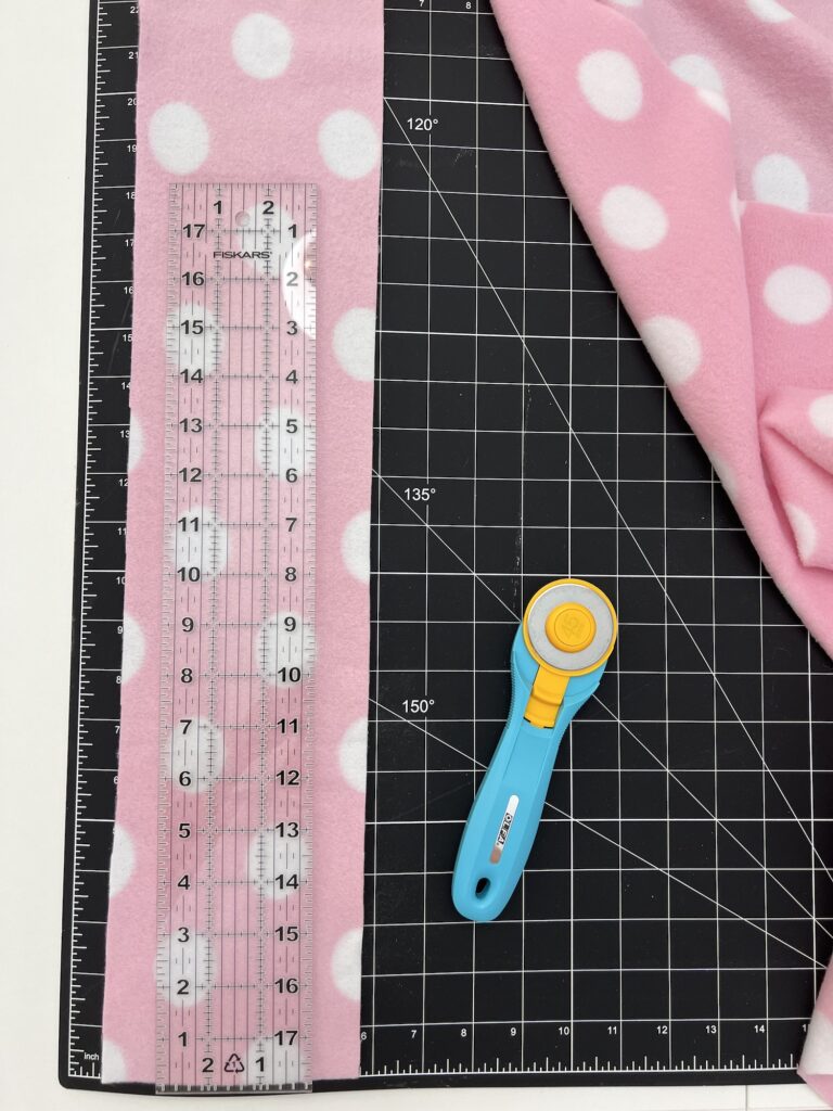 A pink polka dot fabric on a cutting mat with a clear ruler and a blue rotary cutter, perfect for crafting your next cozy fleece headband.