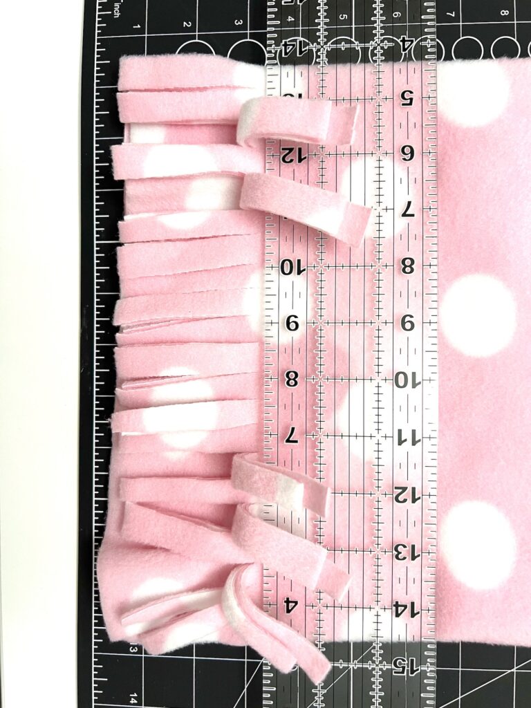 A pink and white polka dot fleece fabric, perfect for crafting a cozy fleece hat, sits with its cut fringes on a cutting mat. A clear ruler is aligned on top, displaying measurements in inches.