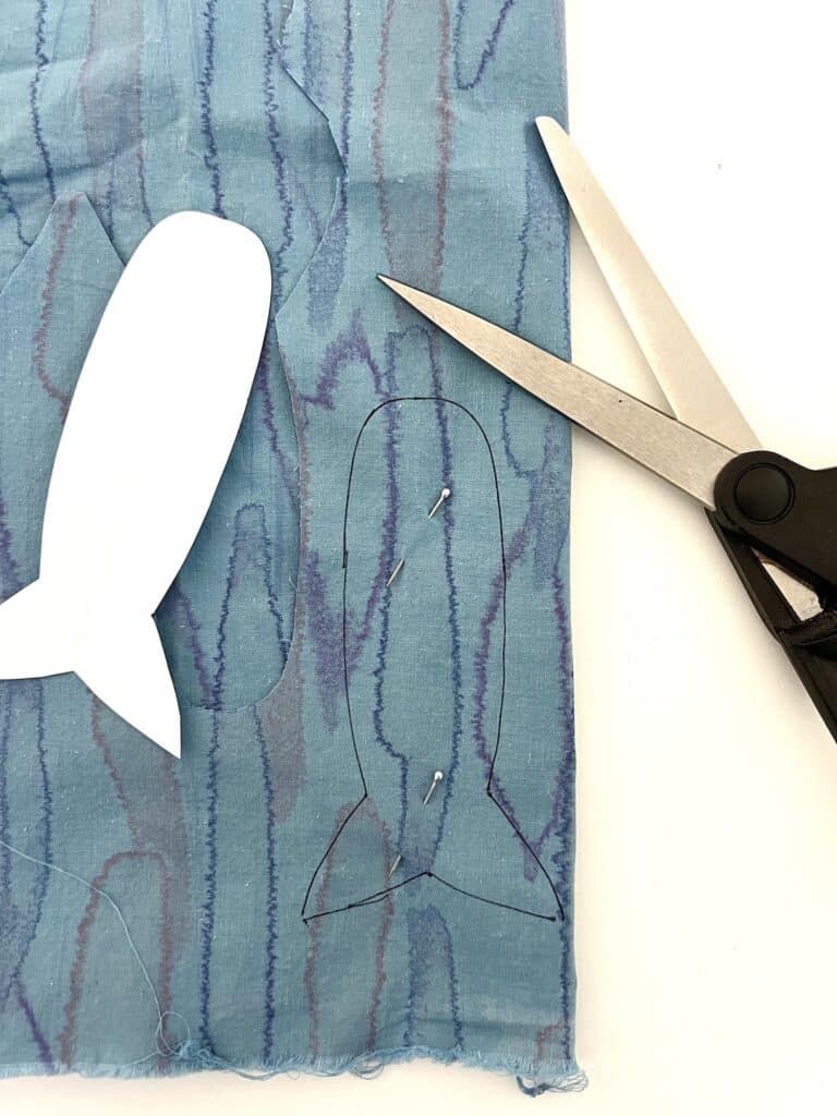 Scissors and a cut-out sardine shape on blue patterned fabric. A paper template is nearby.