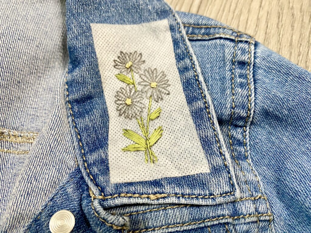 Denim jacket with embroidered floral patch depicting daisies with green leaves on a fabric strip.