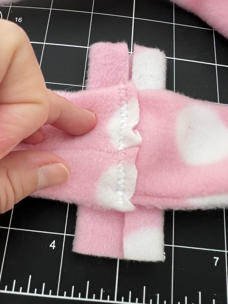 A hand gestures toward a seam on pink fleece fabric with white circles, lying atop a black cutting mat with white gridlines—perfect material for crafting a cozy fleece headband.