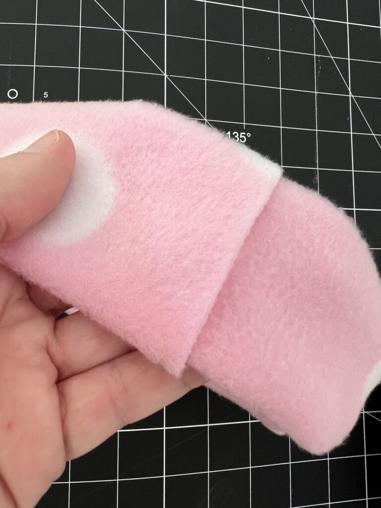 A hand holding a folded piece of pink fleece fabric with a white circle pattern, perfect for crafting a cozy fleece headband, against a black grid cutting mat background.