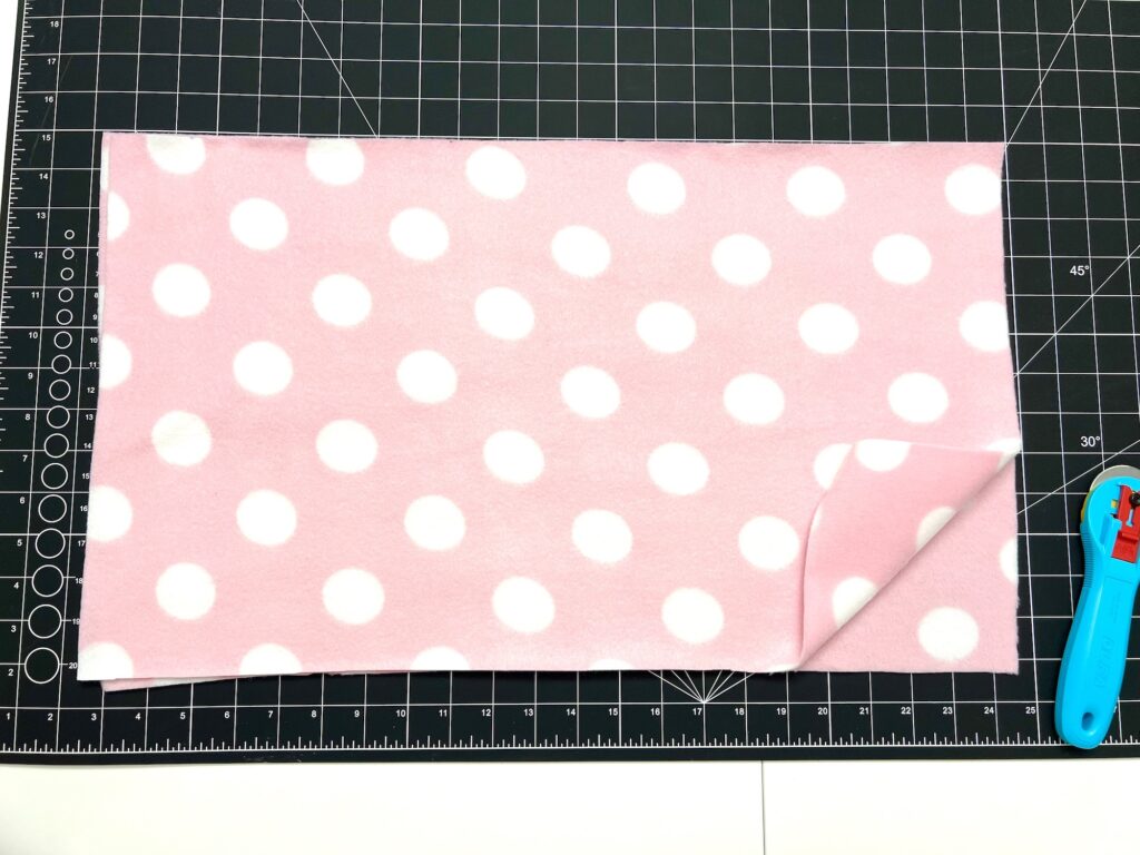 A piece of pink fabric with white polka dots is laid on a black grid cutting mat, ready to be crafted into a cozy fleece hat. A rotary cutter is positioned on the mat's right side, poised to bring the creative vision to life.