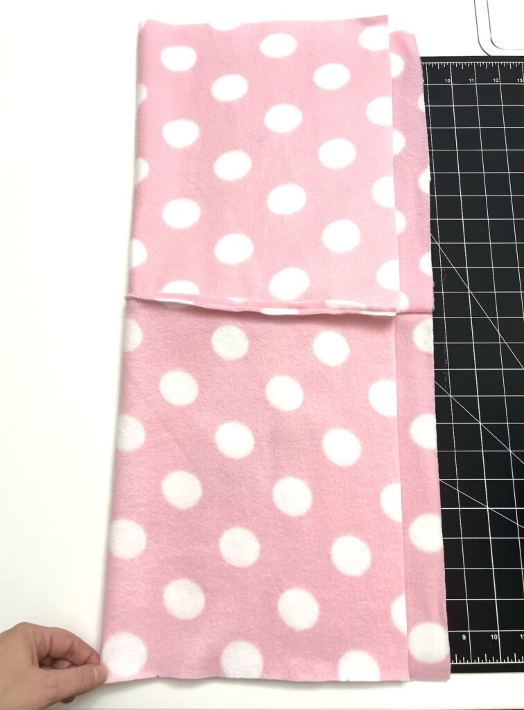 A hand holds the edge of pink fleece fabric with white polka dots, folded neatly on a cutting mat, ready to be transformed into a cozy fleece hat.