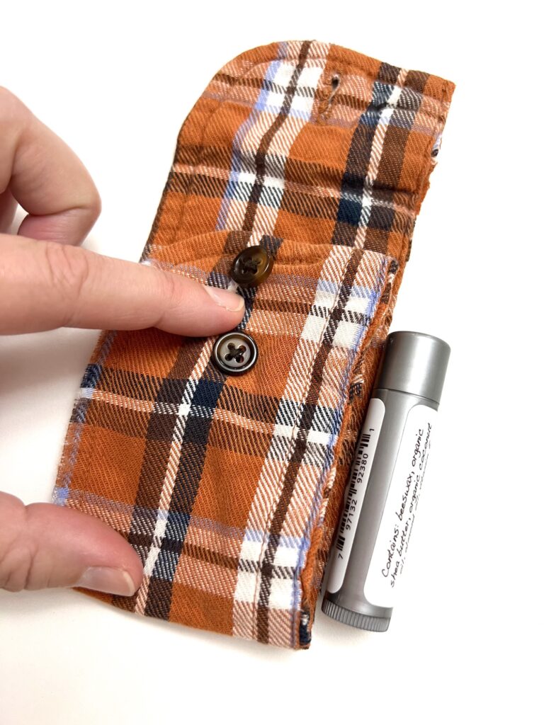 A hand is touching an orange plaid fabric pouch with buttons. A lip balm tube is beside the pouch.