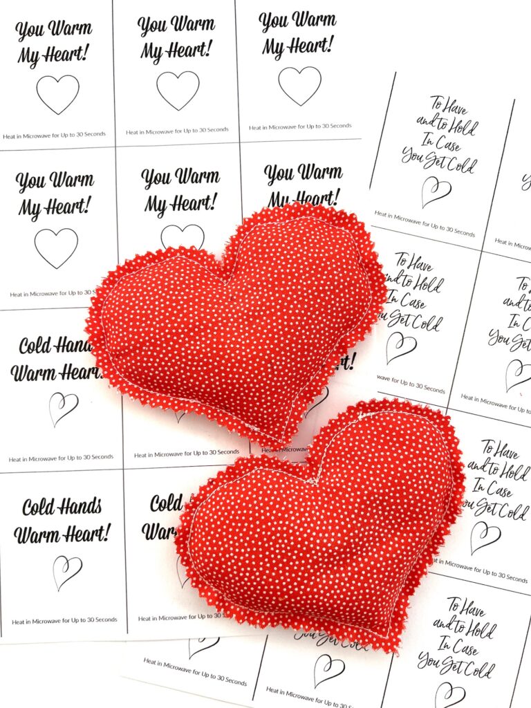 Two red heart-shaped fabric sachets with white dots rest on paper tags that read "You Warm My Heart!" and "Cold Hands Warm Heart," featuring DIY hand warmer heating instructions.