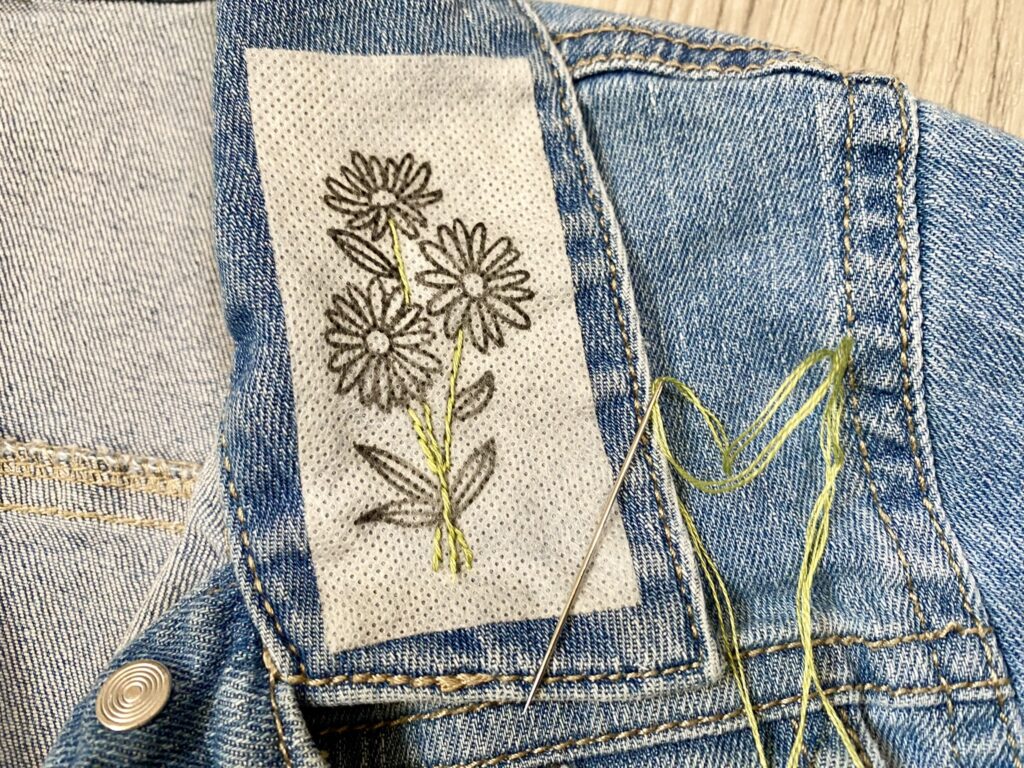 Embroidery thread, needle, and floral design on a denim fabric.