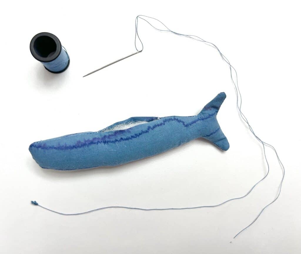 A blue fabric fish figure reminiscent of sardines, complete with a needle and blue thread, sits elegantly against a pristine white background.