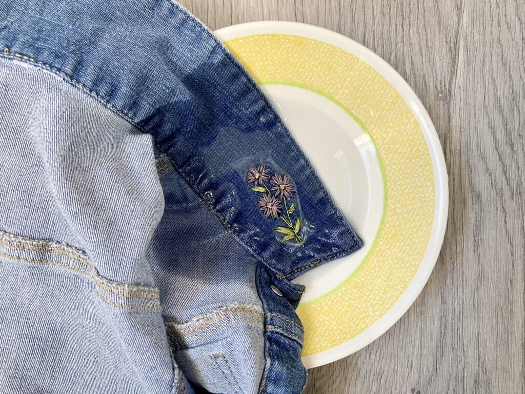 A denim jacket with floral embroidery on the cuff is draped over a white and yellow patterned plate on a wooden surface.