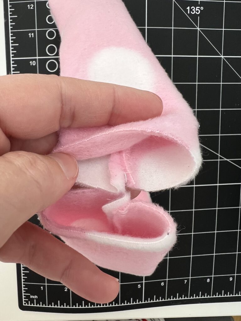 A hand holds a pink fabric piece with white patches, resembling the soft texture of a fleece headband, in front of a black cutting mat with measurements.