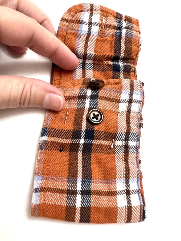 Two fingers hold an orange and black plaid fabric piece with two buttons and a few pins.