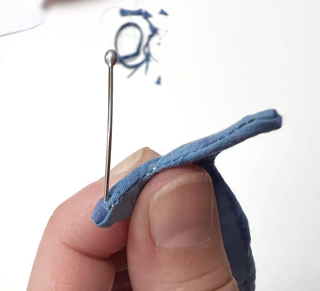 Close-up of a hand delicately holding a piece of blue fabric, with a pin inserted through it, reminiscent of the precision with which sardines are packed into their tin.