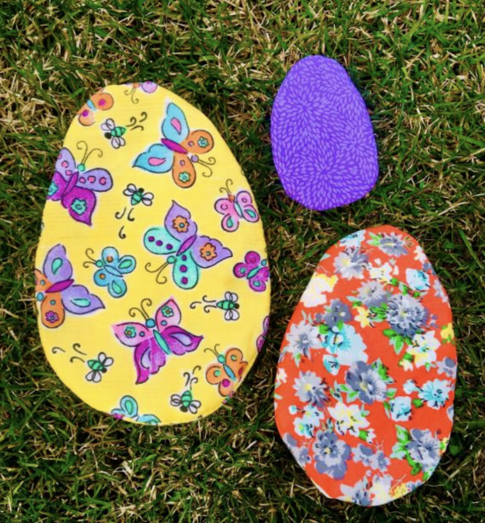 Three colorful, patterned egg shapes on grass: a large yellow egg with butterflies, a smaller purple egg featuring intricate bunny sewing patterns, and a medium orange egg with a floral design.