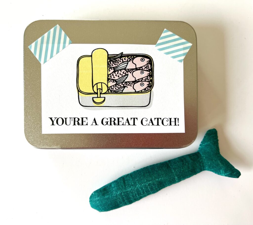 A tin featuring an illustration of sardines and the playful text "You're a great catch!" is adorned with two pieces of tape above the image. Nearby, a green fabric fish adds a touch of whimsy, making this presentation as charming as it is unique.