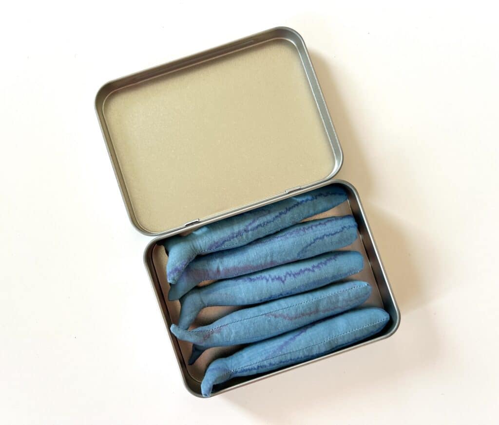 A metal tin resembling a can of sardines contains six plush, blue objects that resemble beans, displayed against a neutral background.