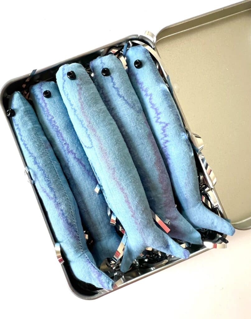 Five plush toy fish with blue bodies and black eyes, snugly arranged in a metal box with a hinged lid, cleverly mimicking a tin of sardines.