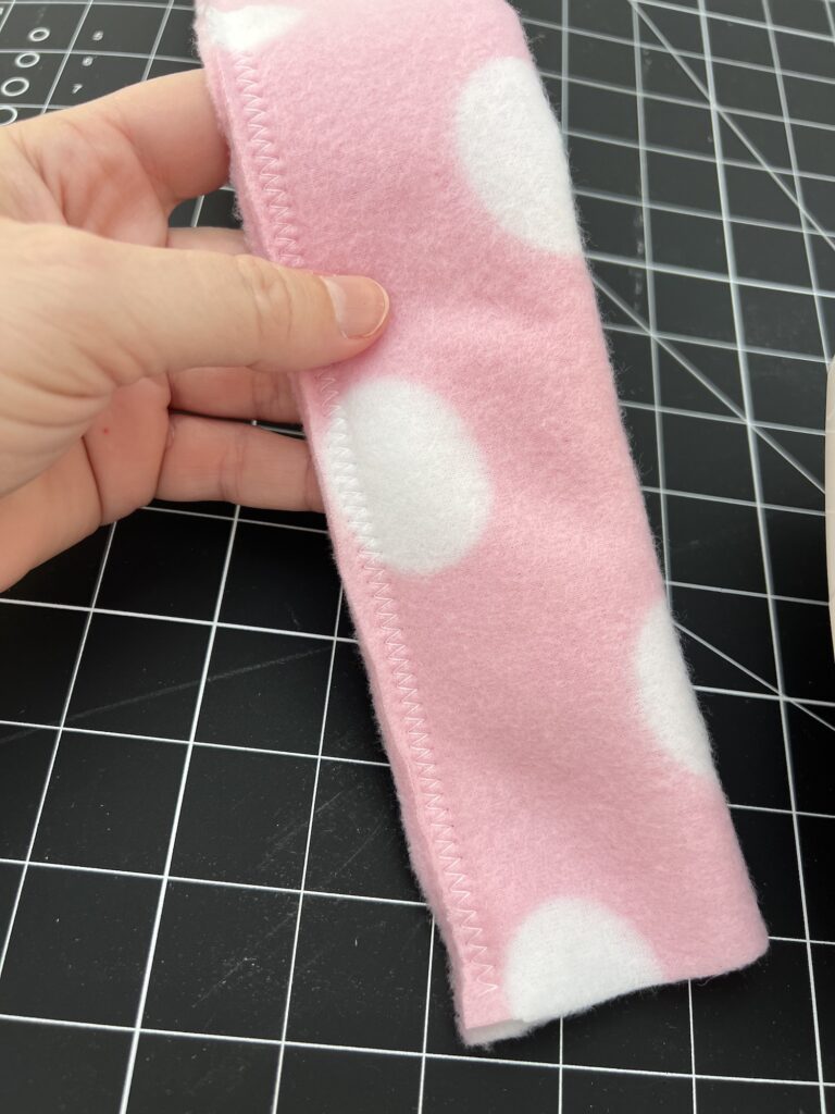 A hand holds a piece of pink fleece fabric with large white polka dots, skillfully stitched at the edges, perfect for crafting a cozy fleece headband. A black grid cutting mat lies in the background.