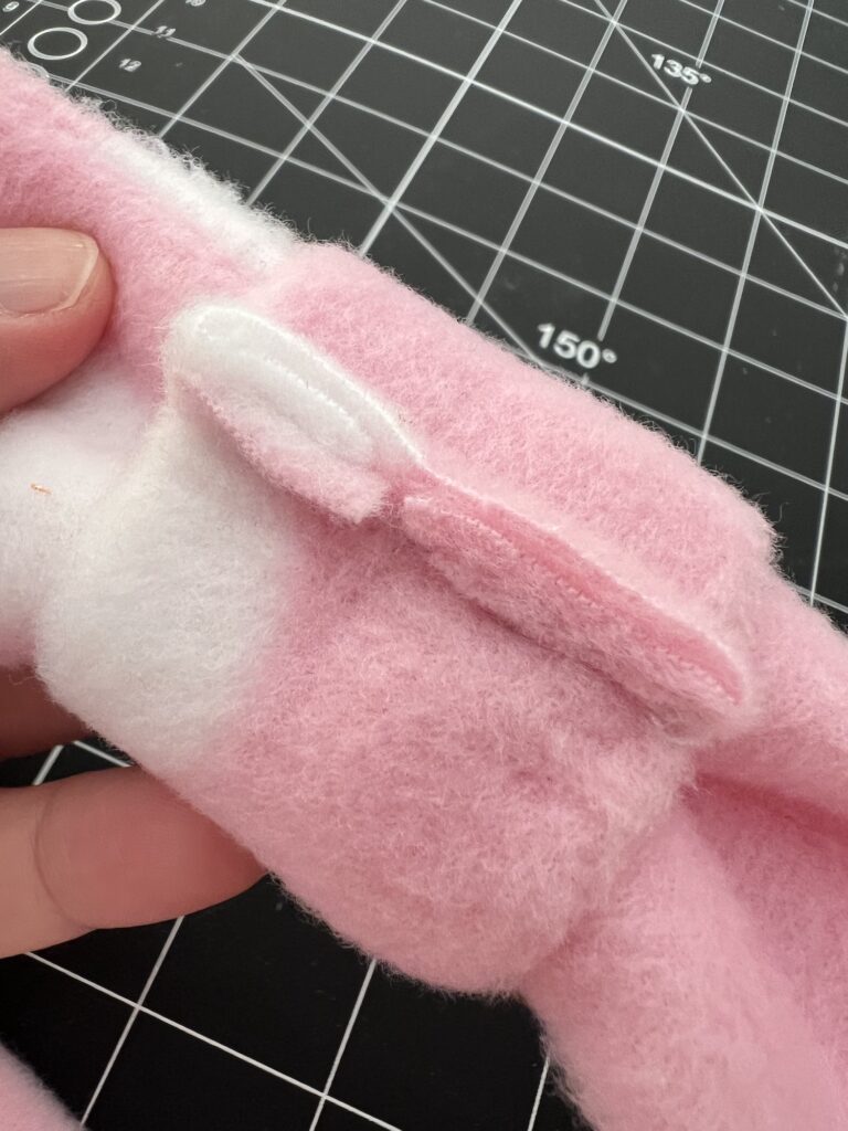 Close-up of a hand holding a pink and white plush fleece headband with a small, sewn ear-like detail, set against the backdrop of a grid cutting mat.