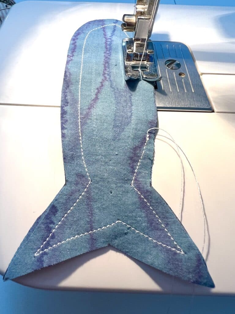 A piece of blue fabric with a fish tail shape is being sewn on a sewing machine, reminiscent of glistening sardines. The stitching is visible along the edges.