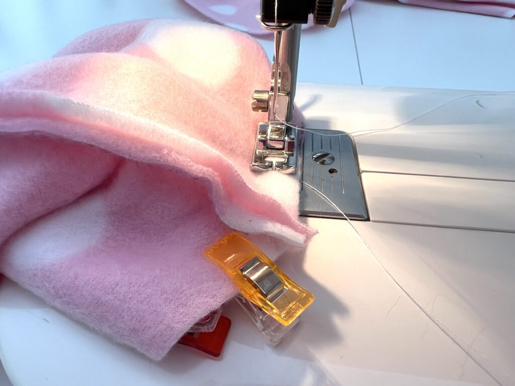 Close-up of pink and white fabric being sewn on a sewing machine. Two fabric clips hold the layers together.