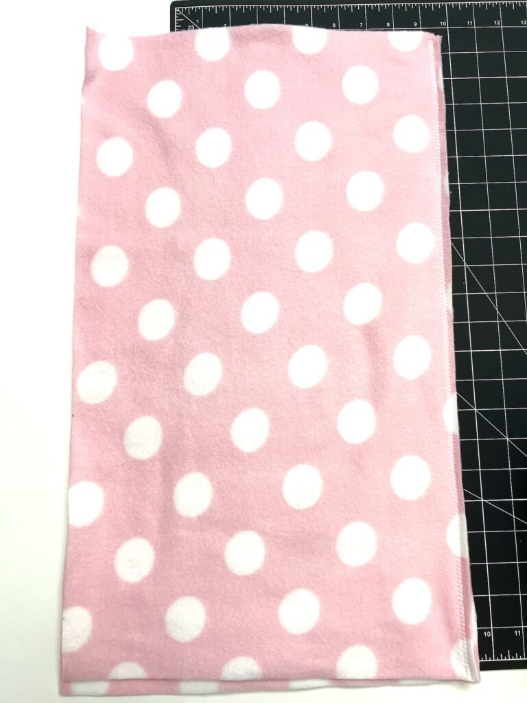 A pink fabric with white polka dots is laid on a black cutting mat with a grid pattern, ready to be transformed into a cozy fleece hat.