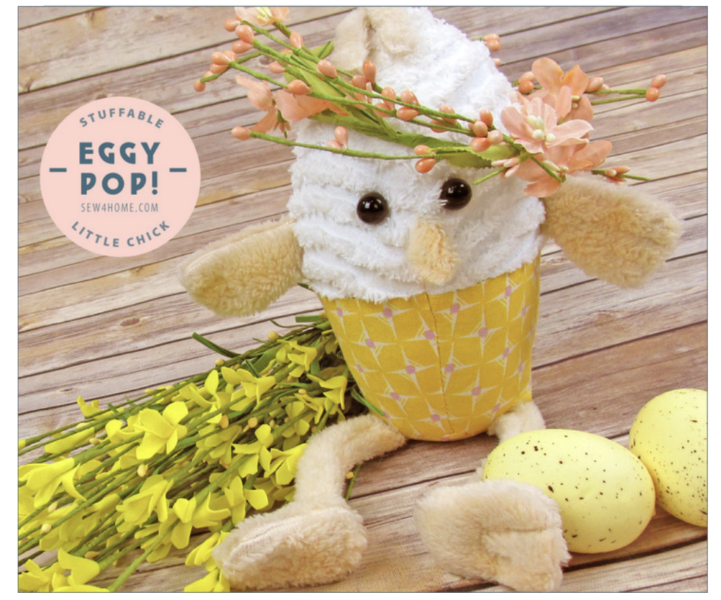 Plush toy chick with a floral crown sits in a yellow-patterned shell cup, surrounded by yellow flowers and spotted eggs. A sign reads "Stuffable Eggy Pop! Little Chick," perfect for spring decor alongside your favorite bunny sewing patterns.