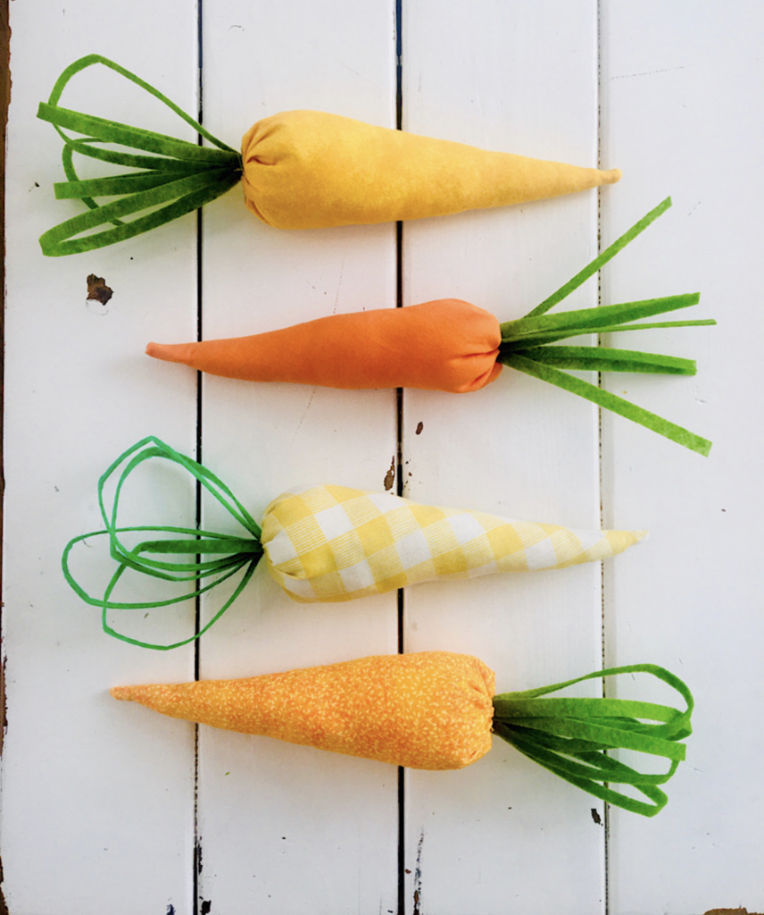 Four fabric carrots in yellow and orange, with green fabric tops, are displayed on a white wooden surface