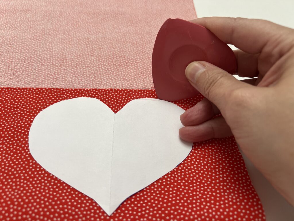 Hand tracing a white paper heart shape on red fabric with a chalk marker, the first step in crafting cozy DIY hand warmers.