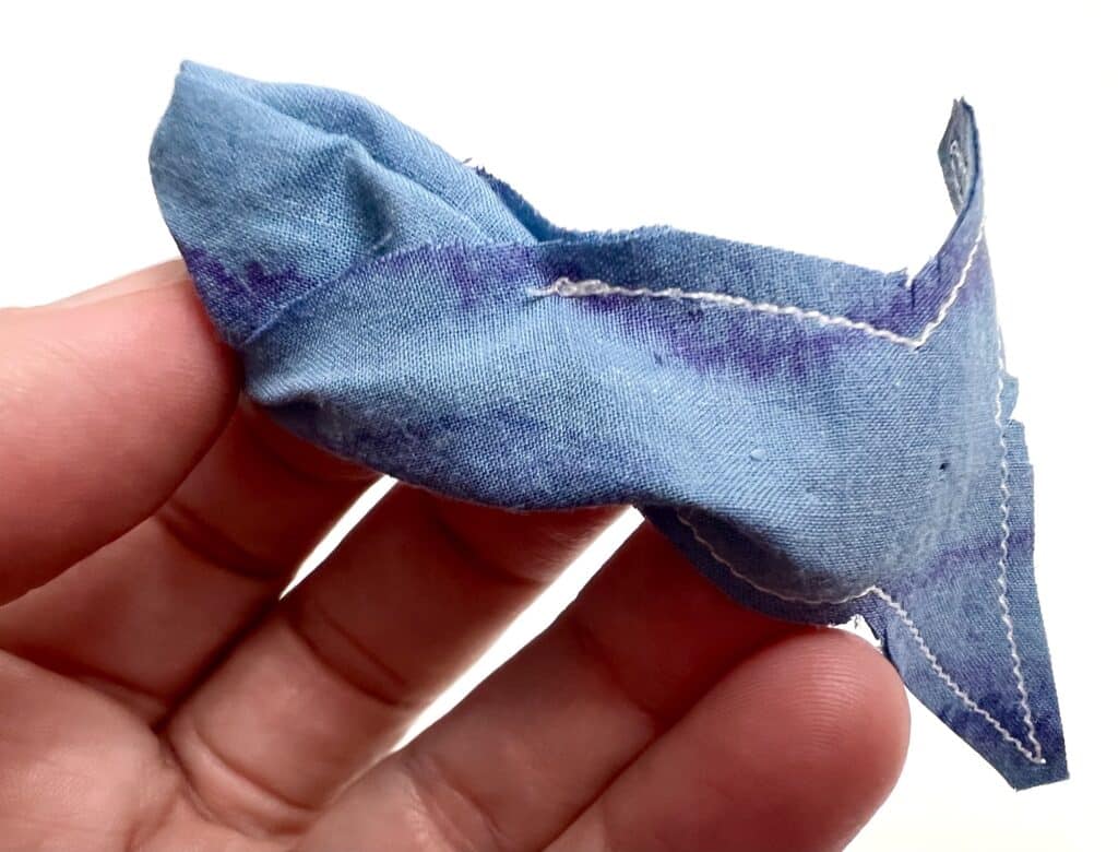 A hand holding a small, folded piece of blue fabric with visible stitching and a purple stain, reminiscent of the unexpected burst of color when sardine juice meets linen.