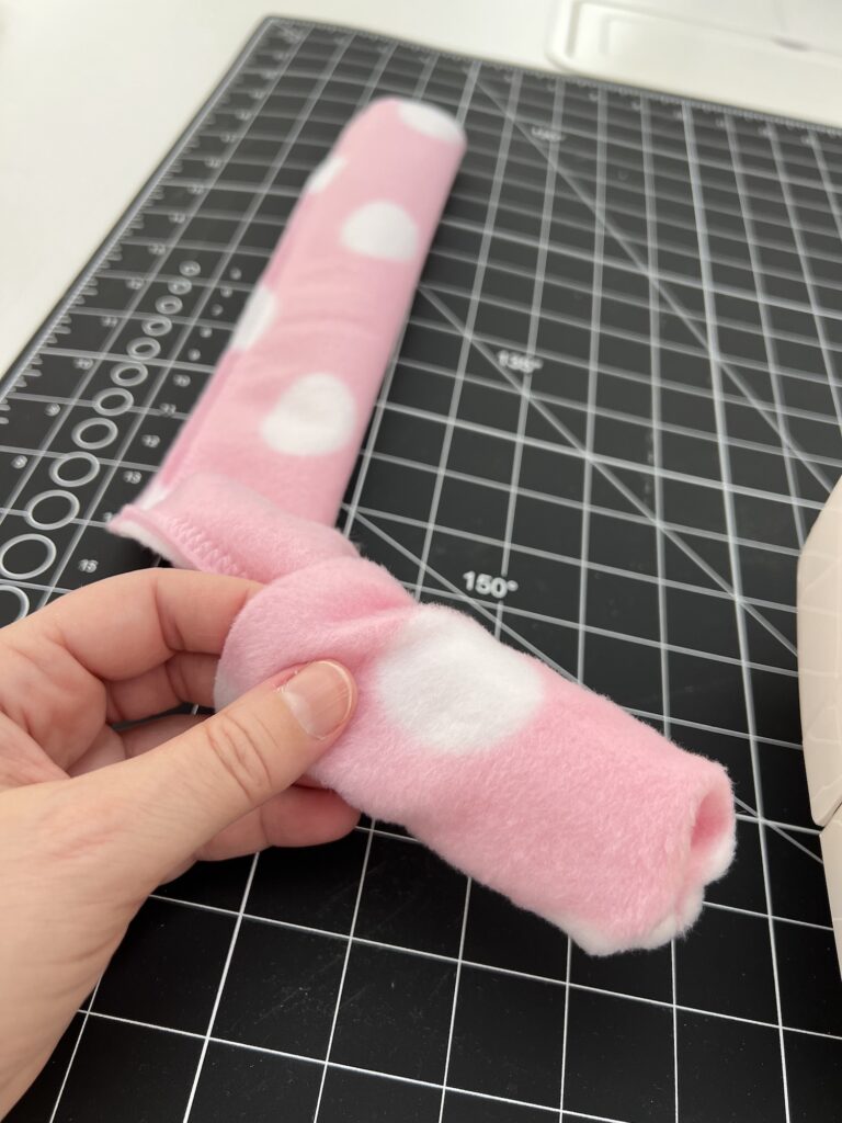 A person holds a partially sewn pink fleece headband with white polka dots on a black cutting mat with grid lines.