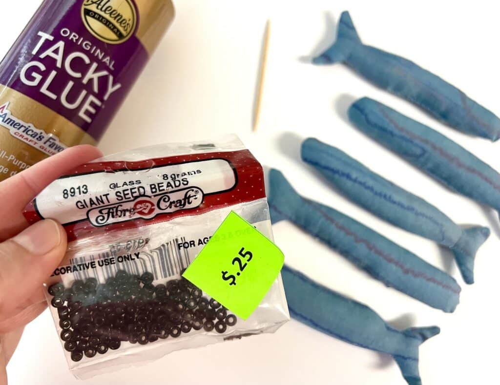 Pack of black glass seed beads with a bottle of tacky glue, a toothpick, and four blue stuffed sardines on a white surface.