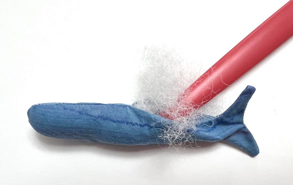 A blue whale toy, reminiscent of sardines in a vast ocean, is ensnared in white netting and placed beside a red plastic straw on a pristine white background.