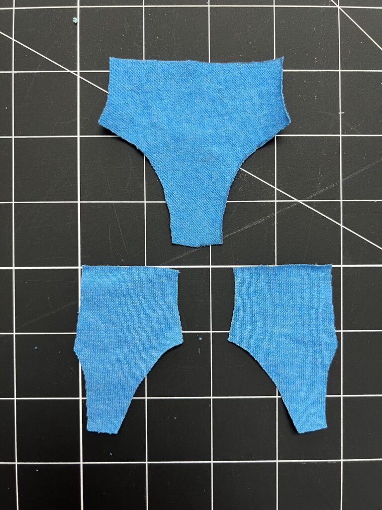 Three small blue fabric pieces are cut in shapes resembling clothing items and placed on a black grid cutting mat.