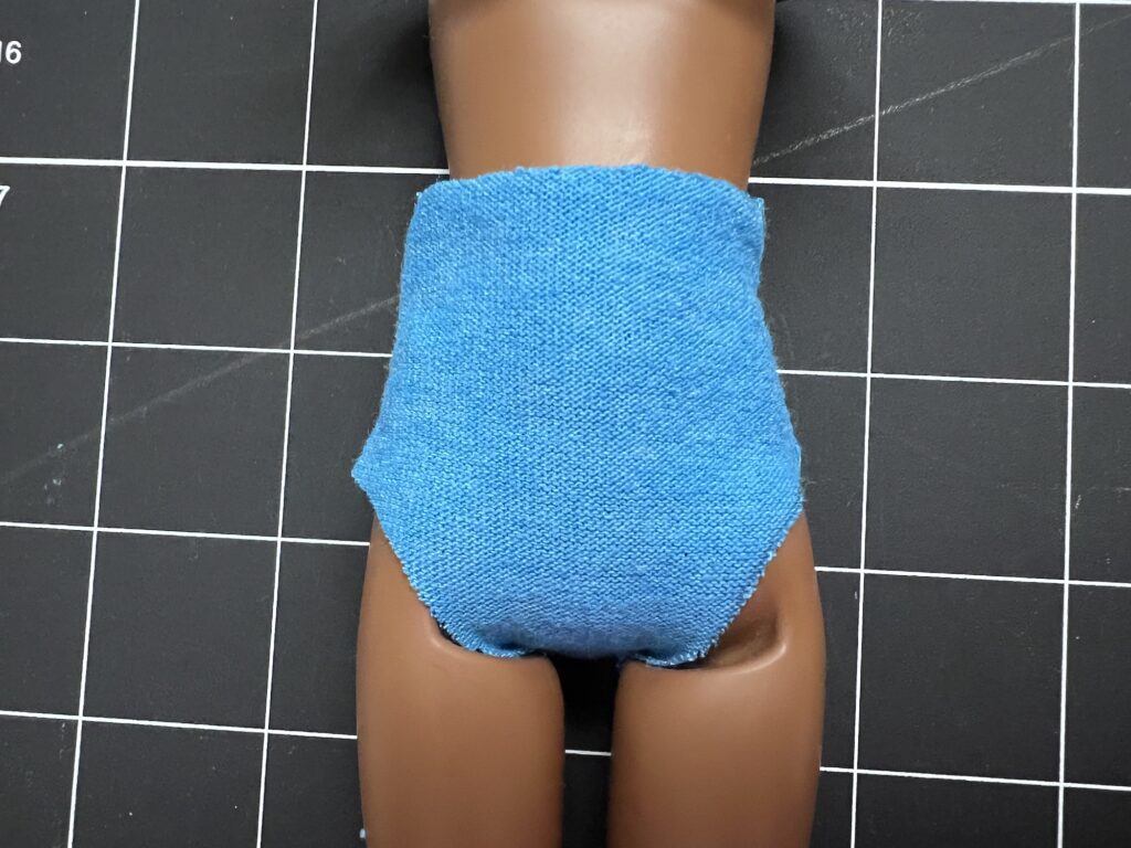 A doll's lower torso and legs are shown wearing a blue fabric covering on a grid-patterned surface.