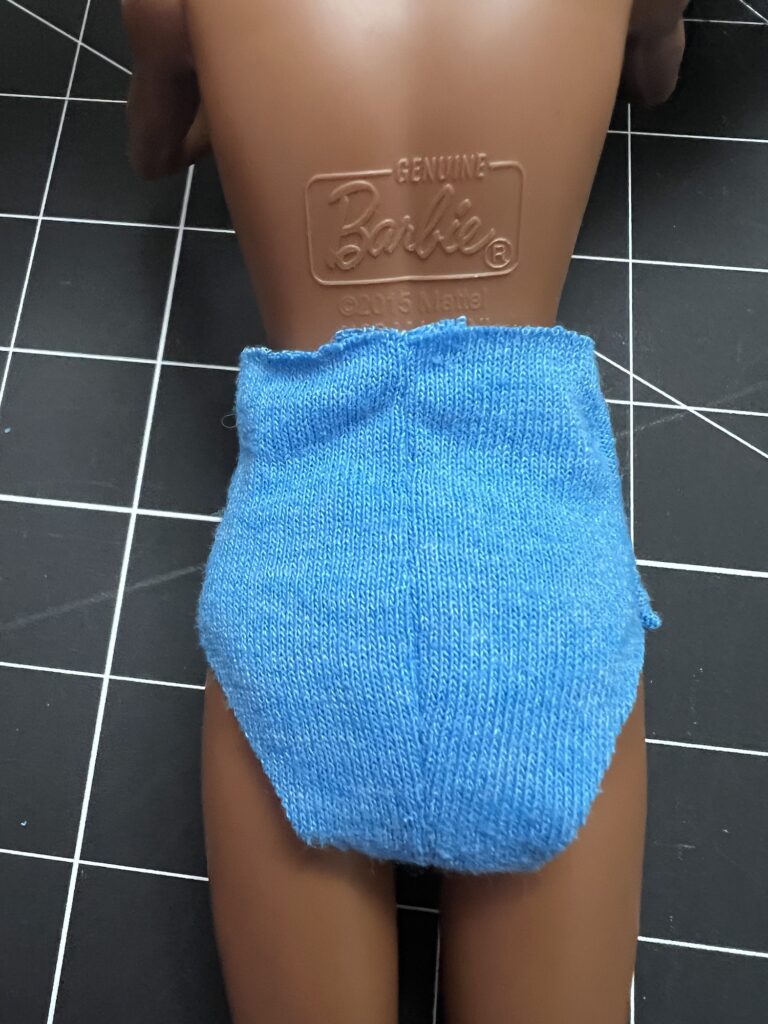 A doll wearing blue knitted shorts lies face down on a grid-lined surface. The word "Barbie" is embossed on the doll's back.