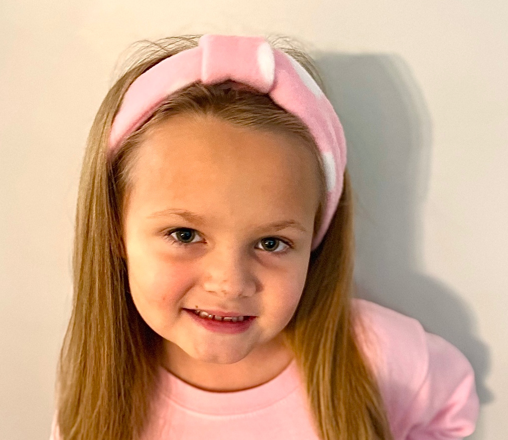 A young girl with long hair and a pink fleece headband, wearing a matching pink top, smiles at the camera against a plain background.