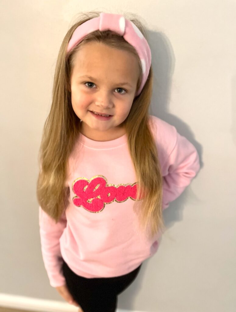 Young girl with long hair wearing a pink fleece headband and a sweater with "Love" written on it, stands in front of a plain wall.