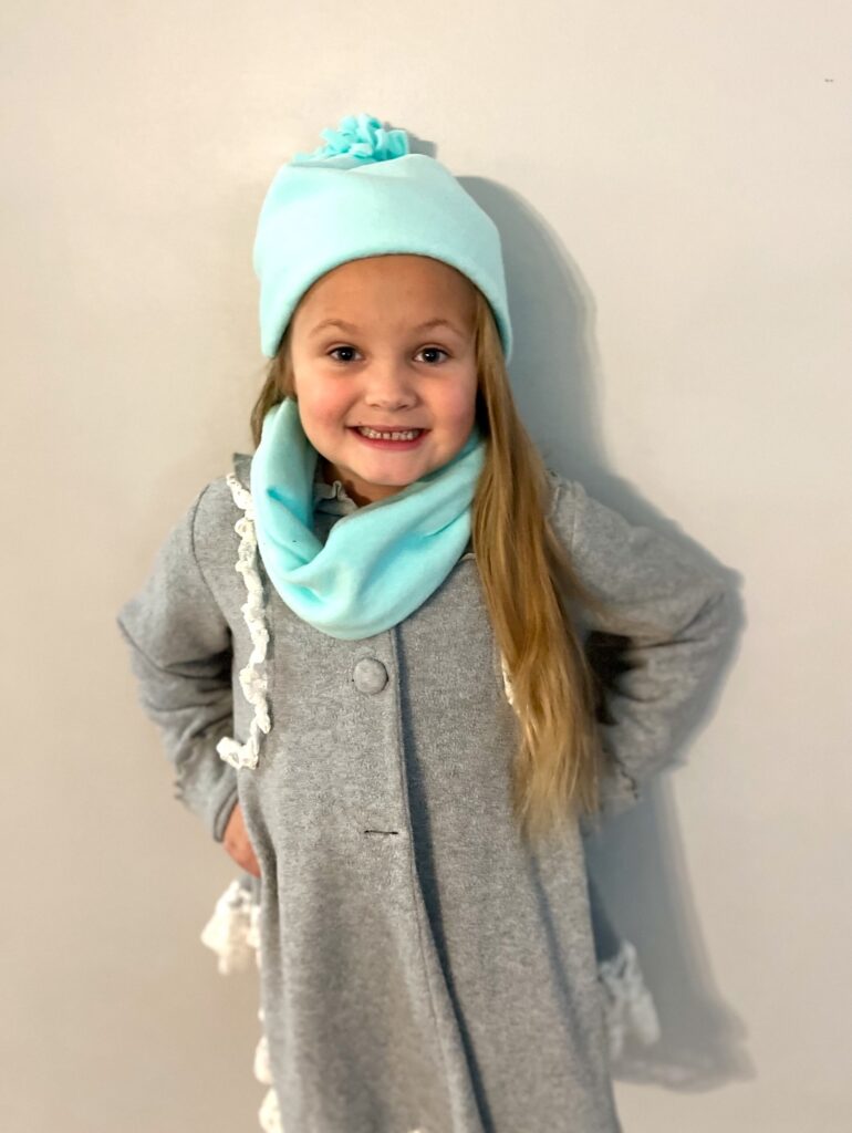 A young child in a gray coat, wearing a light blue fleece hat and matching scarf, stands against a plain background.