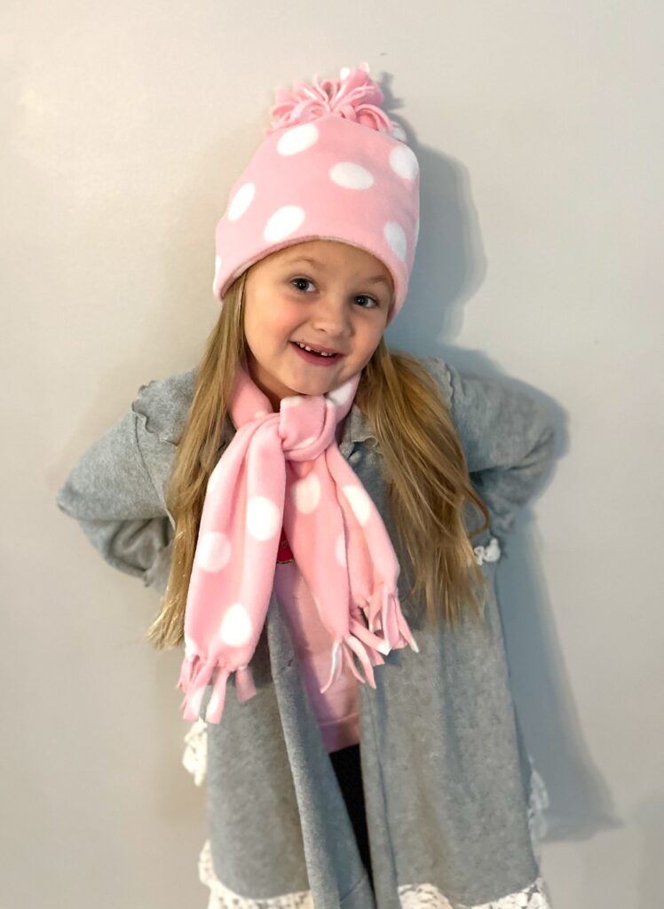 A young girl smiles, donning a pink polka dot fleece hat and scarf, perfectly paired with her gray coat against a plain background.