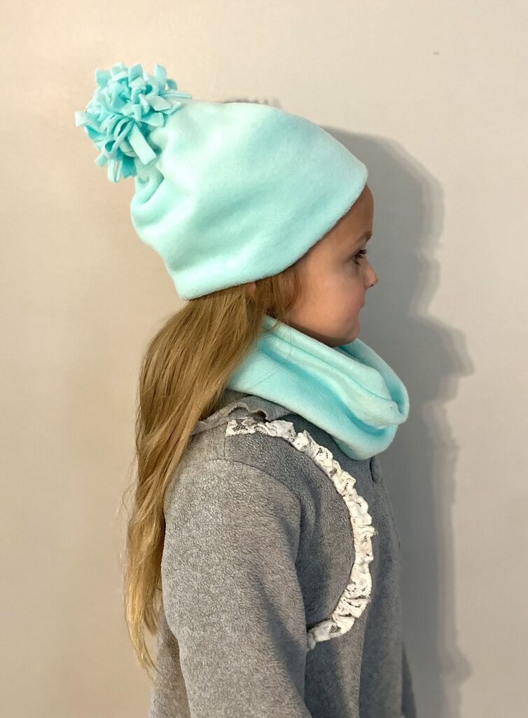 A child in profile wears a light blue beanie with a pom-pom and matching scarf, paired with a grey top. The cozy fleece hat adds an extra layer of warmth against the plain background.