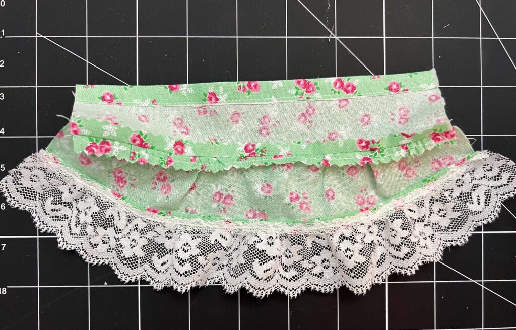 A piece of green floral fabric with pink flowers, perfect for crafting a tiered skirt, features a delicate lace trim and is laid out on a grid cutting mat.