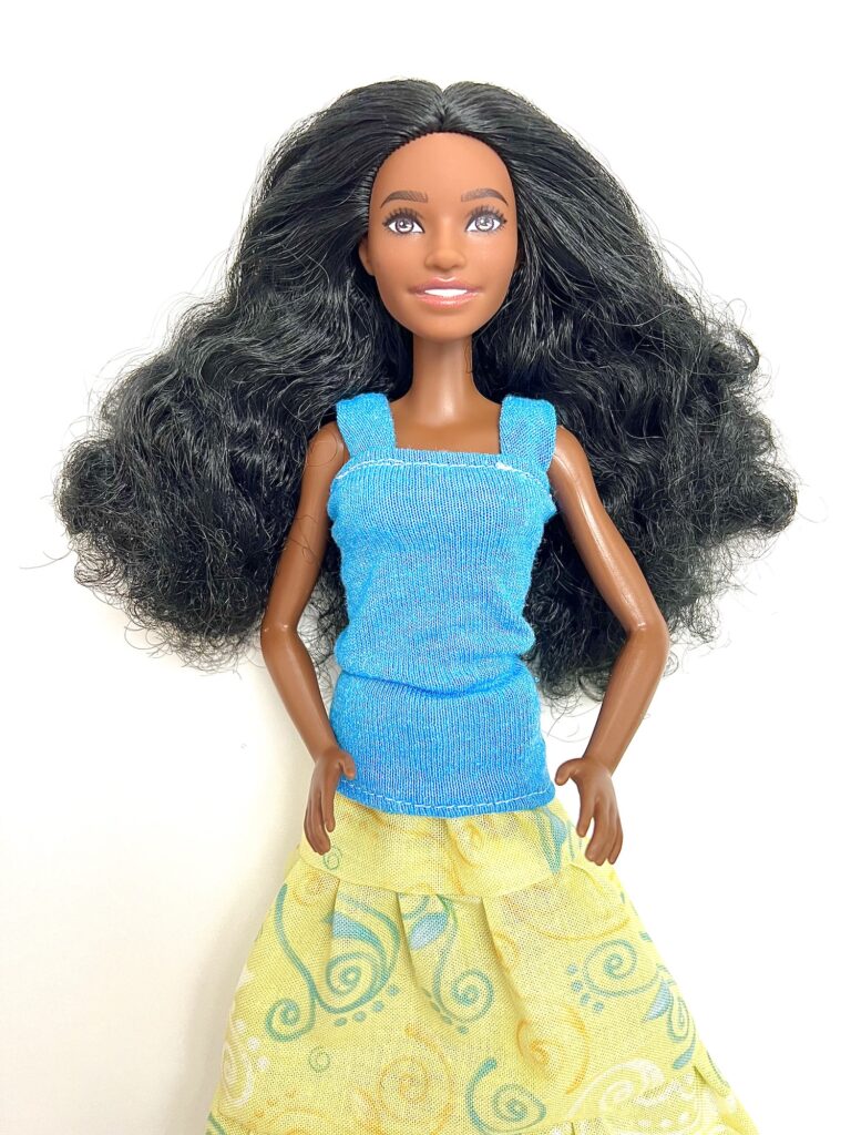 A doll with long black hair, wearing a blue top and a yellow skirt with green swirl patterns, is posed against a plain white background.