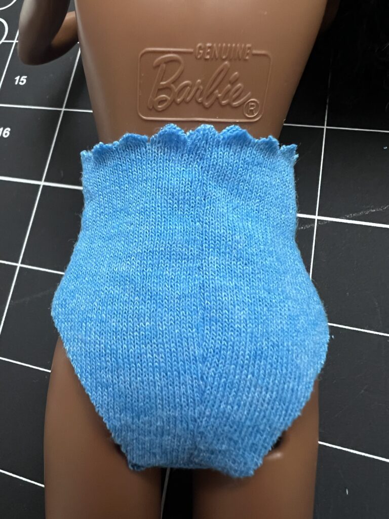 A Barbie doll torso with "Genuine Barbie" marking on the back is wearing a pair of blue knitted shorts.