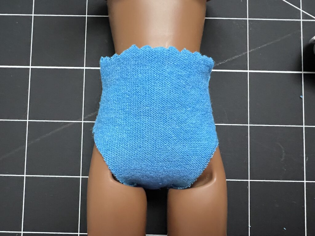 A doll's torso wearing a blue denim-looking garment is positioned on a black grid surface.