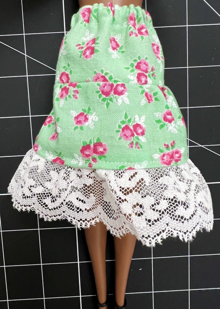 A doll wearing a green floral, tiered skirt with white lace trim is displayed on a gridded black surface.