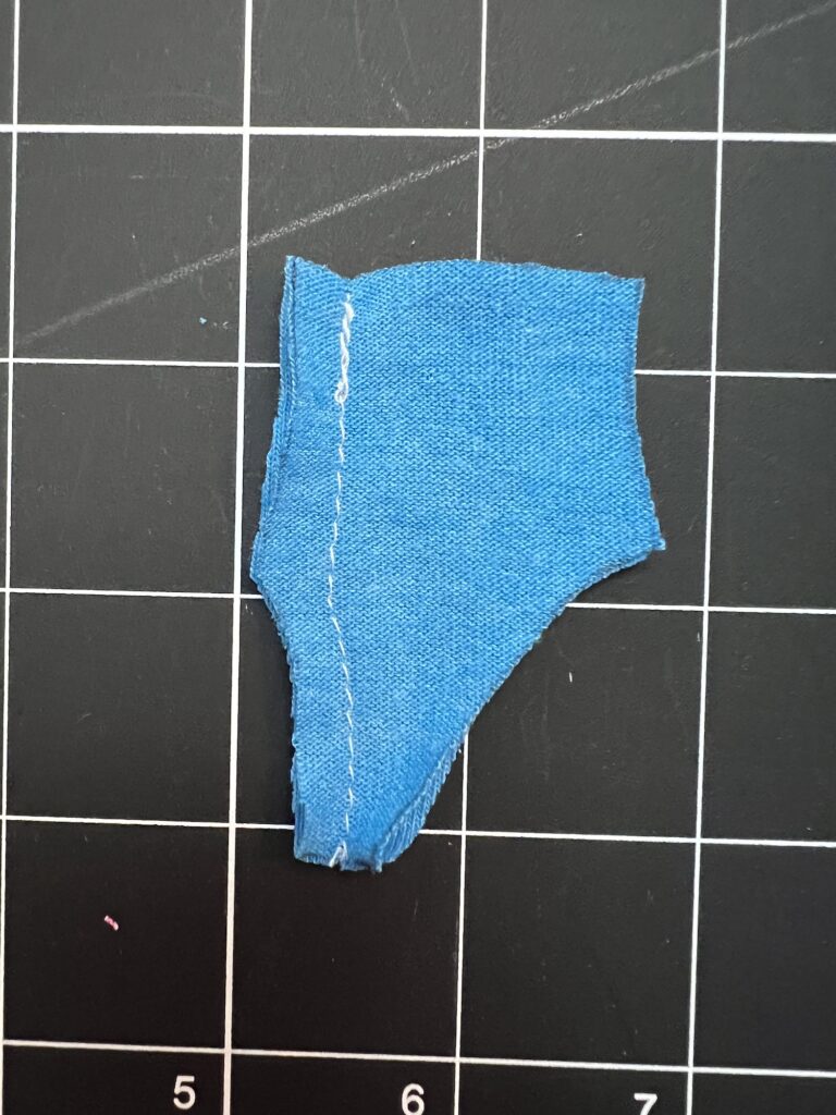 A small, irregularly-shaped piece of blue fabric with a stitched seam, placed on a black grid cutting mat.