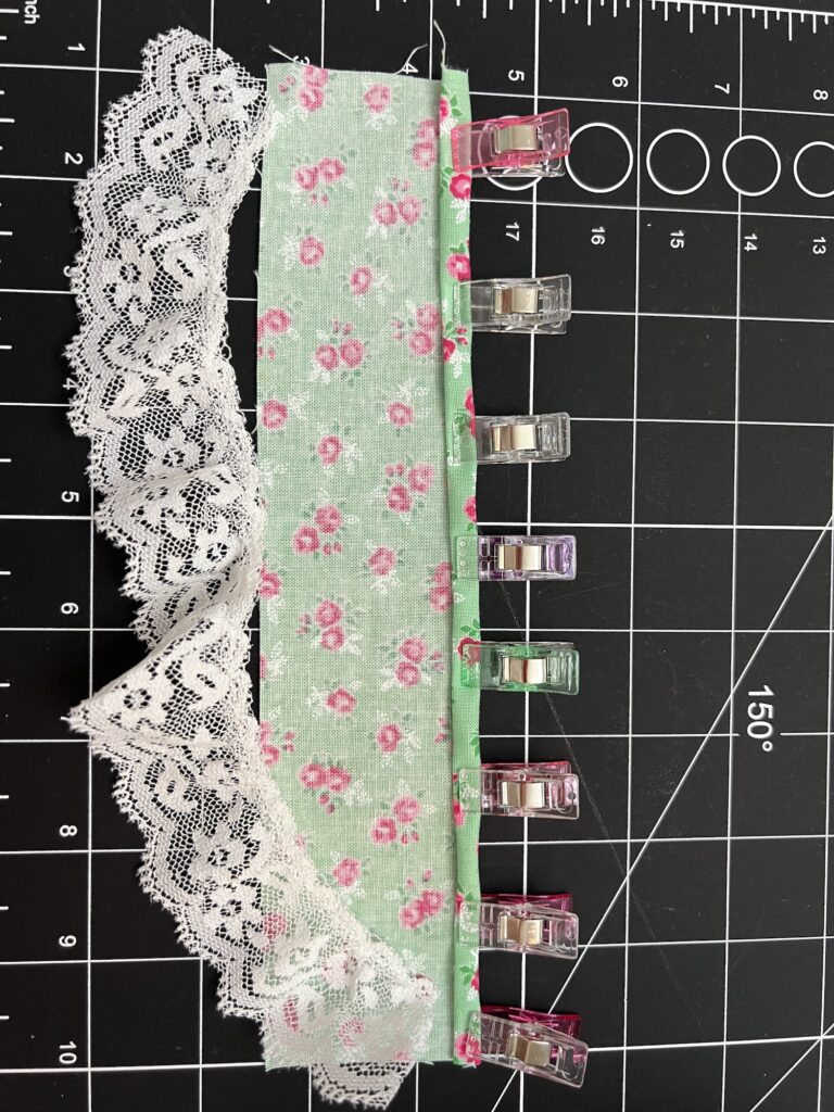 A tiered skirt fabric in green floral with lace trim is neatly secured by multicolored clips on a black cutting mat with measurement markings.