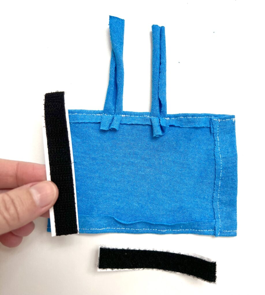 A hand holds a piece of black Velcro next to a blue fabric piece with straps, with another Velcro strip placed below on a white background.